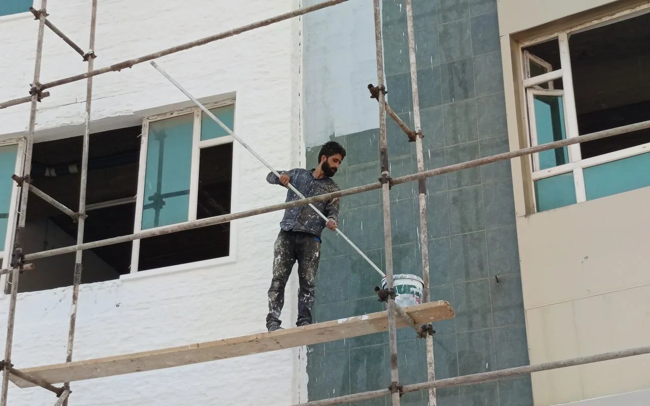 A bearded painter stands on scaffolding using a long-handled roller to paint a building's exterior wall, with a paint bucket nearby.