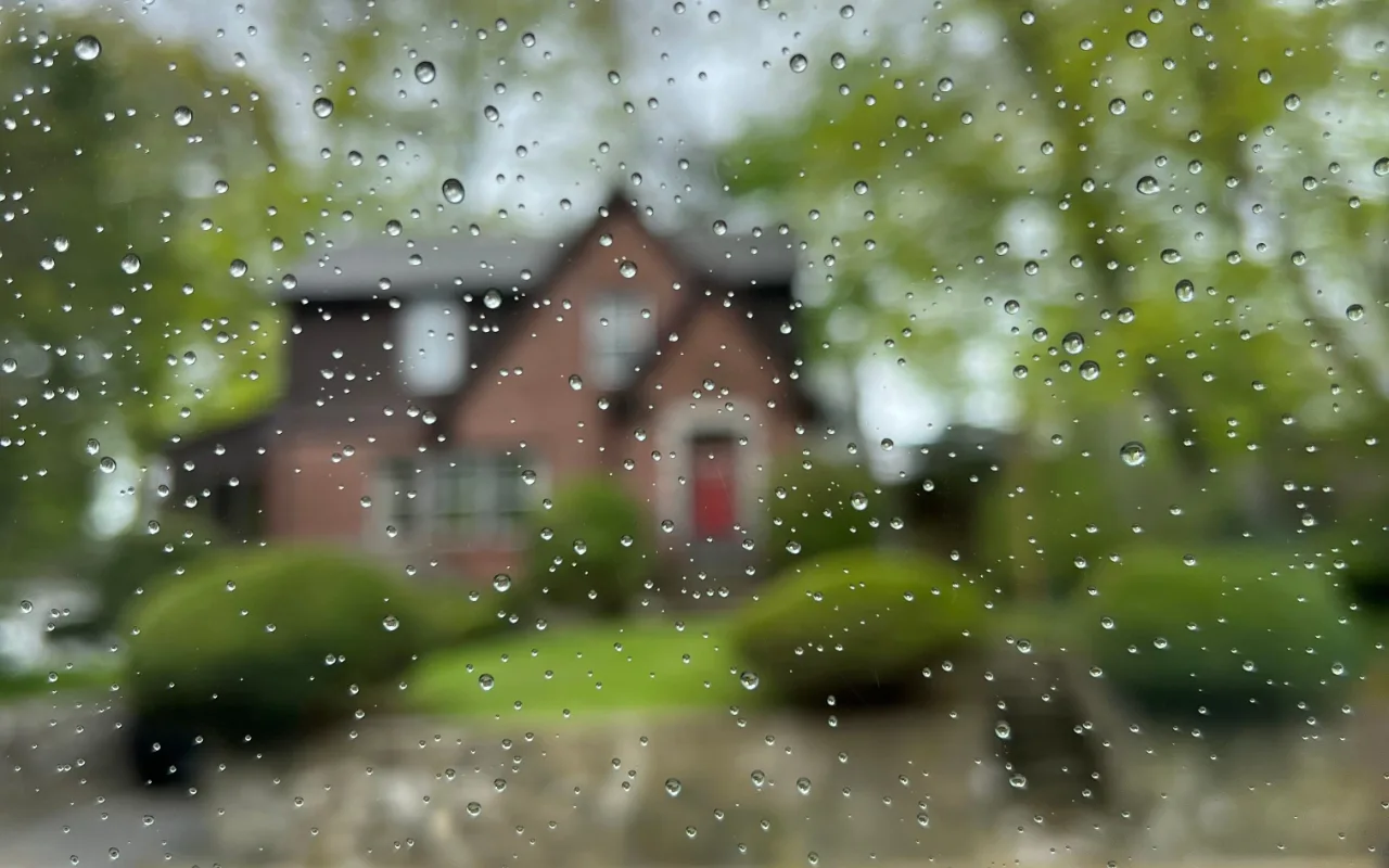 Raindrops on a window pane with a blurred red-doored brick house and lush green shrubs visible beyond.