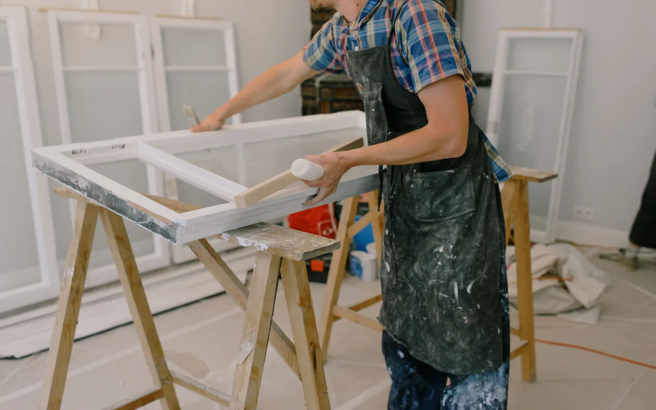 A painter in a paint-splattered apron applies white paint to a window frame resting on wooden sawhorses in a workshop.