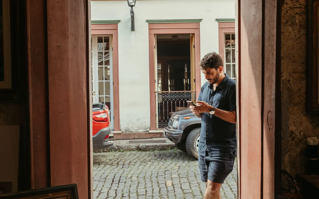 A man in a dark blue shirt and shorts checks his phone while standing in a cobblestone courtyard beside parked vehicles.