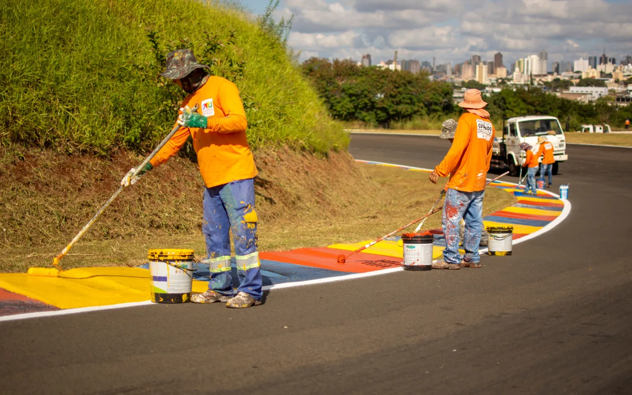 Workers in orange uniforms paint colorful stripes along a curved racetrack curb using rollers and buckets of red, yellow, and blue paint.