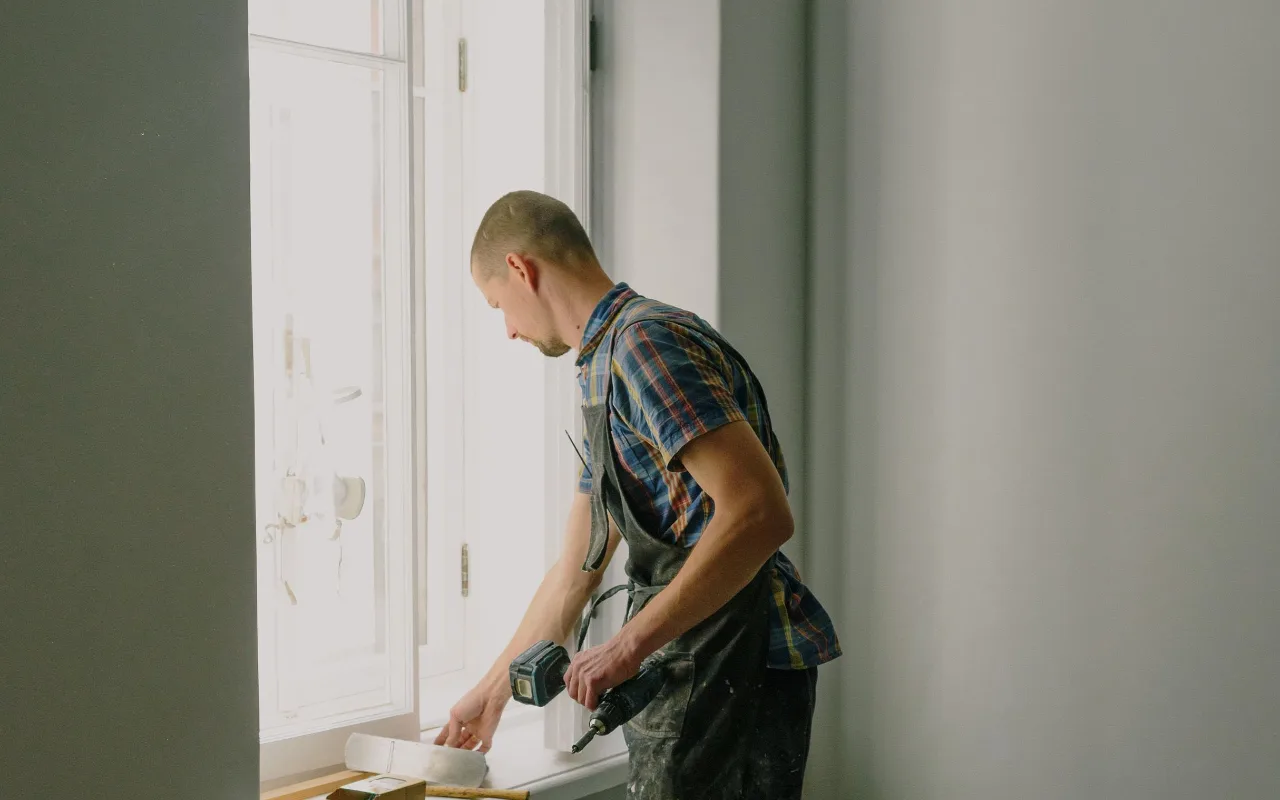 A tradesperson in a plaid shirt and work apron uses a power drill near a bright white window during an interior renovation.