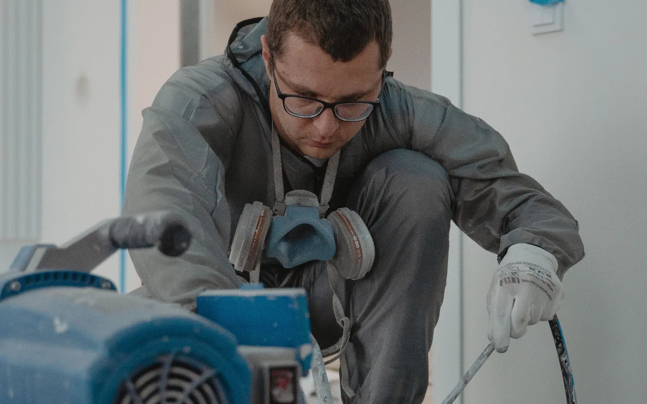 A painter in a gray coverall and respirator mask crouches beside a blue airless sprayer, adjusting a hose while wearing protective gloves.