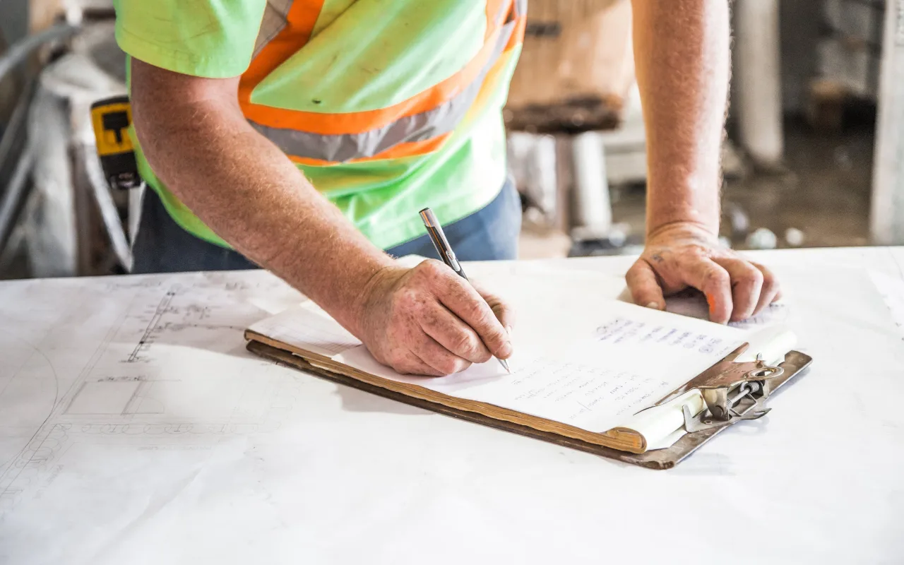 A construction worker in a hi-vis vest writes notes on a clipboard over building blueprints at a job site.