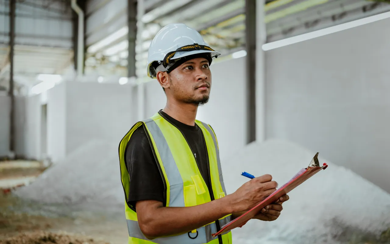 A worker in safety vest and hard hat writes on a clipboard inside a large building, mirroring the inspection and documentation work the argument says wins the job.