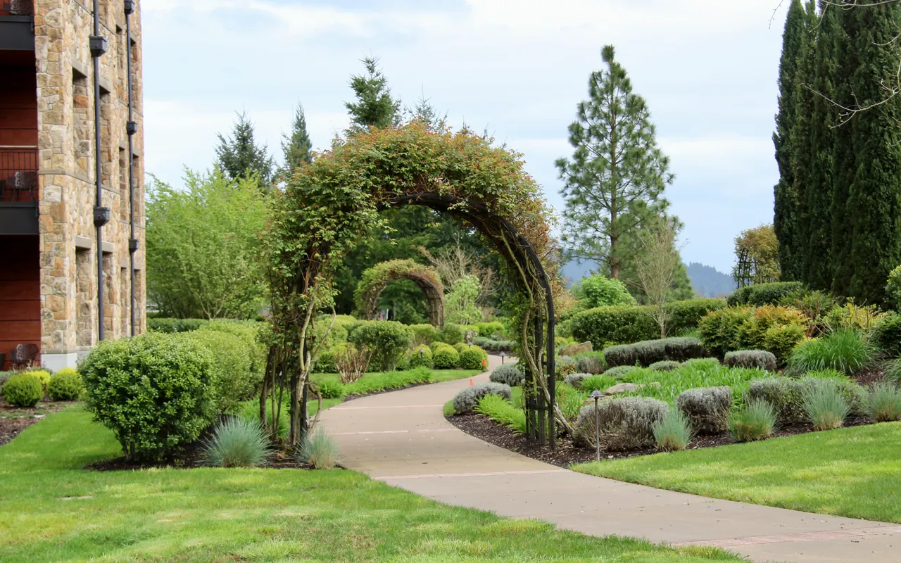 A manicured garden path with rose arches winds past a multi-unit residential building, evoking the ongoing maintenance care that a multi-year program delivers.
