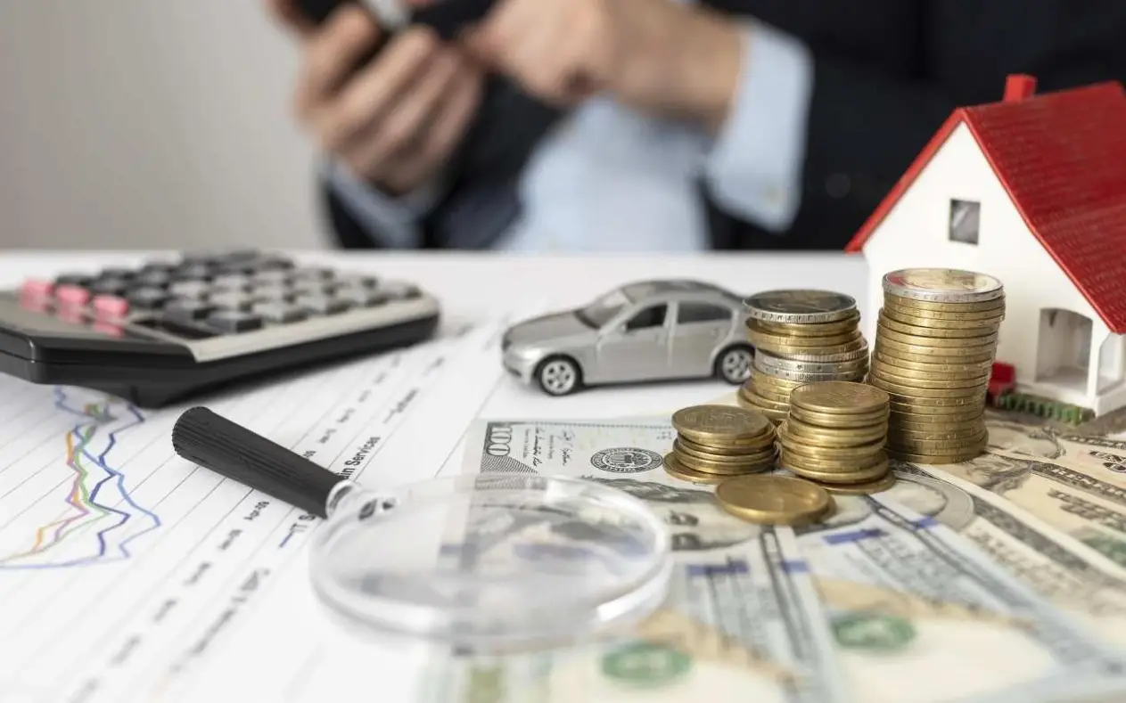 Novčanice, kovanice, kalkulator, maketa kuće i automobila na stolu uz grafikone – prikaz finansijskog planiranja i procene imovine.