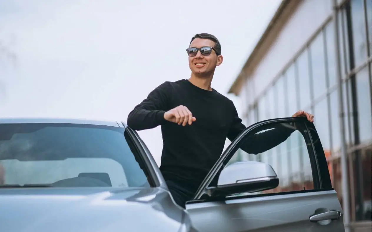 Muškarac sa naočarima u crnom džemperu nasmejan izlazi iz automobila