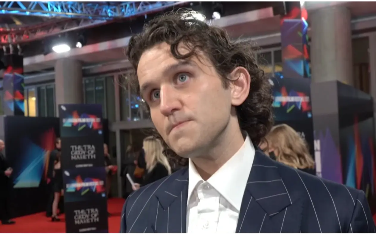 Harry Melling u teget odelu na pruge i beloj košulji sa zamišljenim izrazom lica tokom davanja intervjua