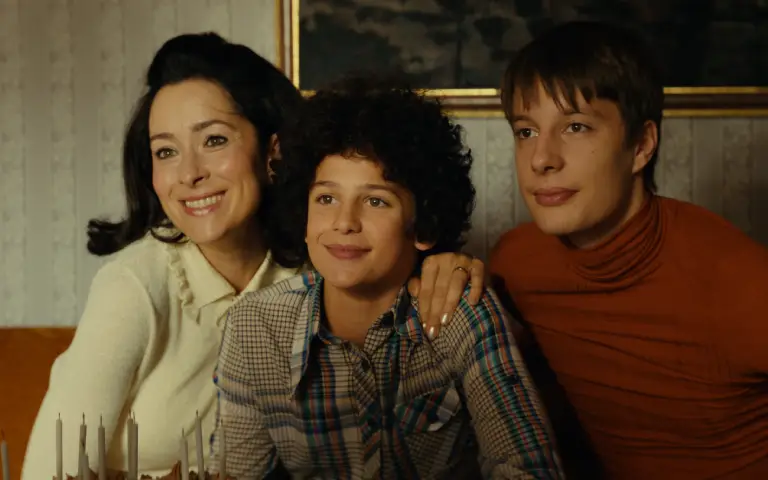 orodična scena iz biografskog filma "Dražen" Topao porodičan trenutak sa nasmejanom ženom, dečakom sa kovrdžavom kosom i tinejdžerom, koji zajedno sede u retro-stilizovanom dnevnom boravku.