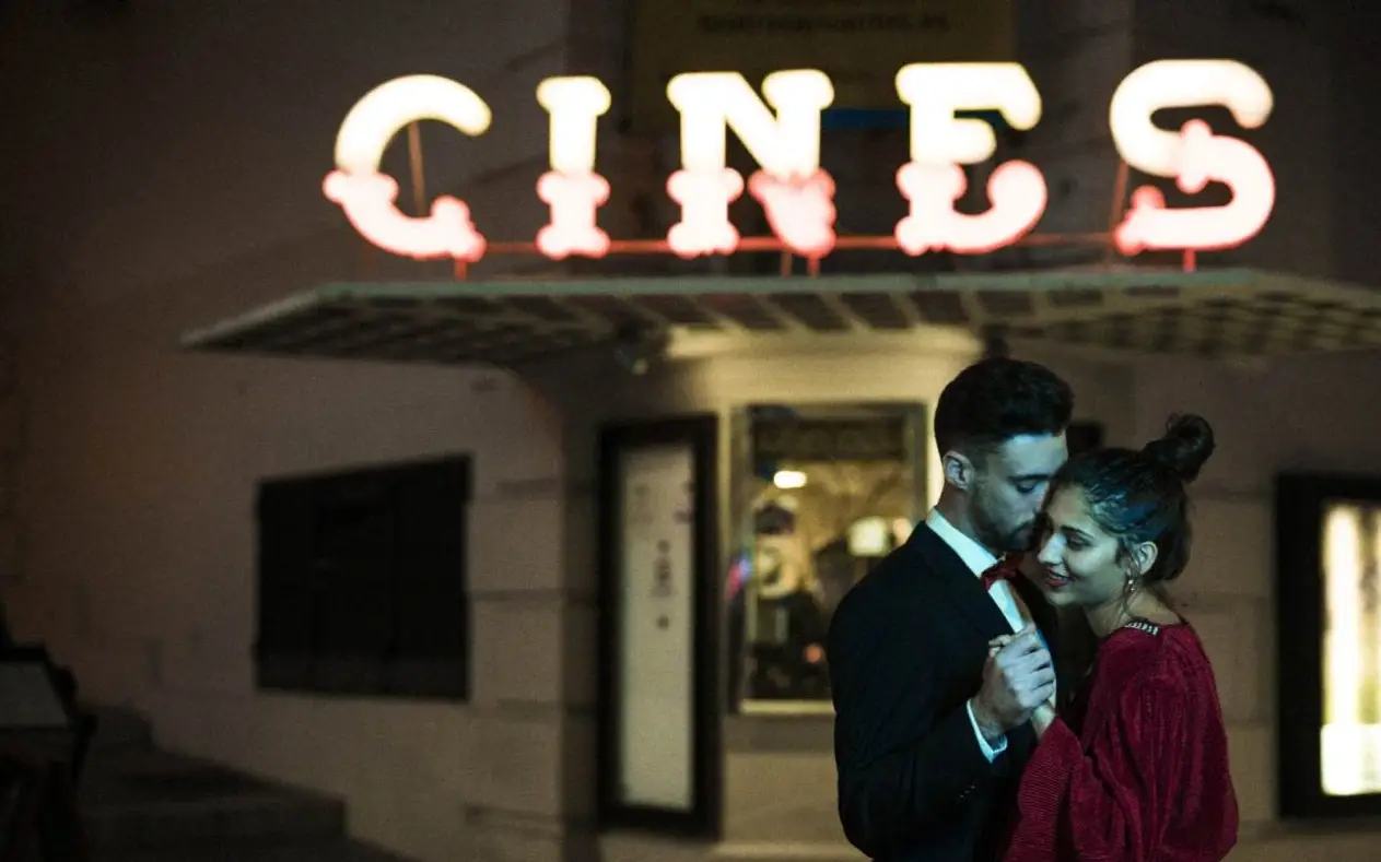 Romantični par zagrljen ispred bioskopa pod svetlima grada, simbol večne ljubavi na filmu.