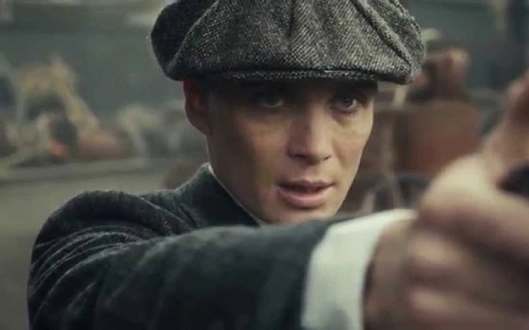  Tommy Shelby, lik iz serije 'Peaky Blinders', u krupnom planu, nosi kapu i drži pištolj uperen ispred sebe