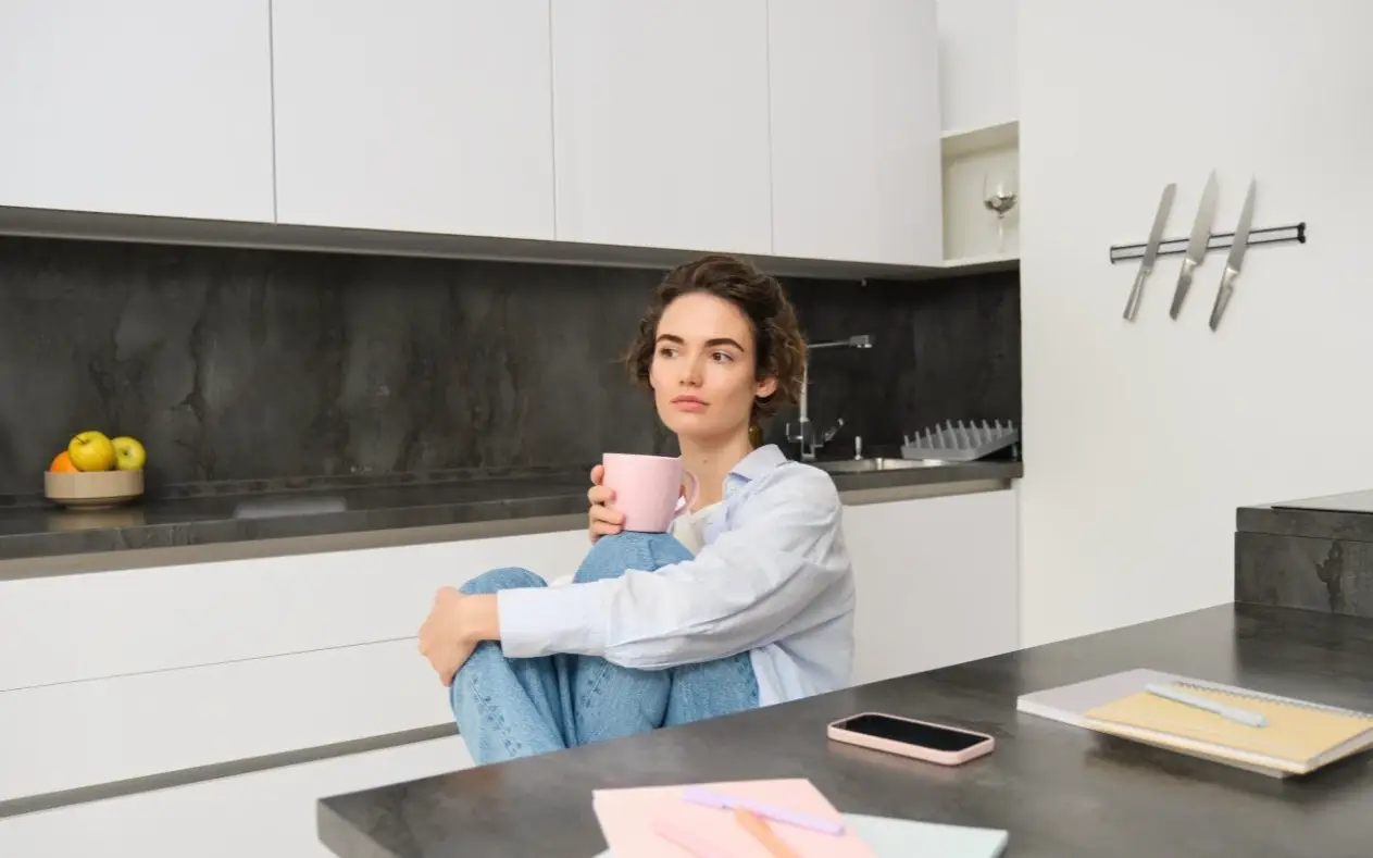 Žena zamišljenog pogleda naslonjena na šolju kafe, u kuhinji u rano jutro