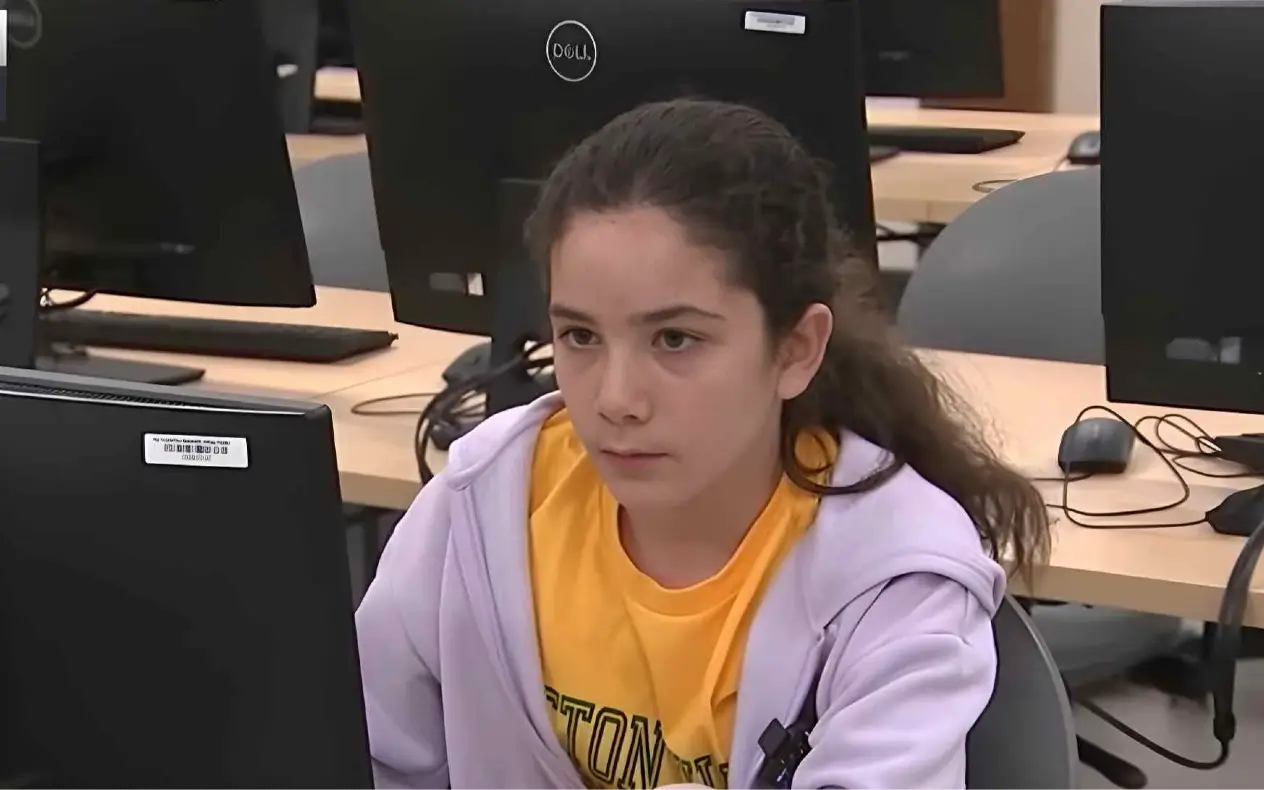 Mlada devojčica u žutoj majici i ljubičastom duksu koncentrisano gleda u monitor dok radi na računaru u učionici opremljenoj sa više računara. U pozadini se vide prazne stolice i monitore marke Dell.