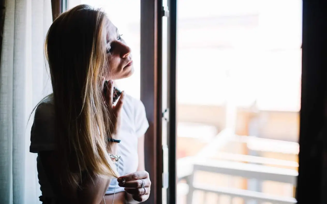 Silueta žene koja stoji kraj prozora i gleda u daljinu, u toplom svetlu zalaska.