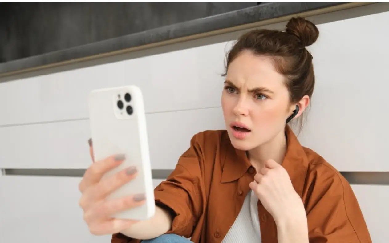 Zabrinuta žena sedi sama u sobi i gleda u svoj mobilni telefon, izraza sumnje i emotivne distanciranosti.
