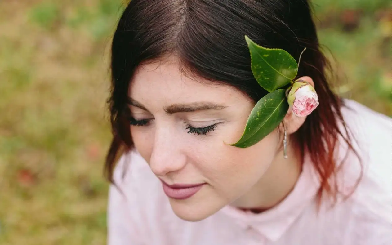 Žena zatvorenih očiju, blagog osmeha, sedi na travi, među cvećem, sa nežnim svetlom koje pada na lice.