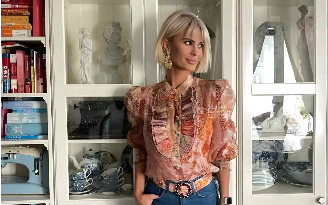 Žena sa kratkom plavom kosom nosi romantičnu bluzu sa puf rukavima i bogatim dezenom u toplim bojama, uparenu sa klasičnim farmerkama i upečatljivim kaišem. Pozira ispred vitrine sa knjigama, antikvitetima i porcelanom, stvarajući spoj sofisticiranog stila i umetničke atmosfere.