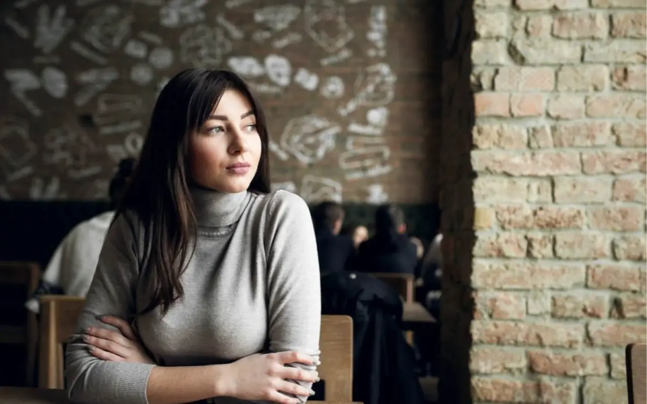 Žena u četrdesetim sedi sama uz prozor, nasmejana ali zamišljena – balans snage i usamljenosti.