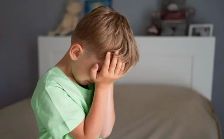 Tužno malo dete, sedi na krevetu, lice prekrivenoo rukama