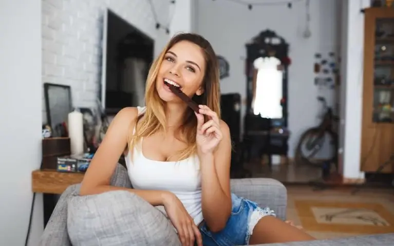 Žena sa osmehom na licu jede čokoladu dok sedi na sivom kauču u dnevnoj sobi