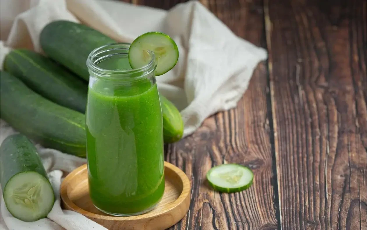 Detoks voda sa krastavcem, limunom, đumbirom i cimetom u staklenom bokalu na letnjem stolu Stakleni bokal sa aromatizovanom detoks vodom, kriškama krastavca, limuna, jabuke, svežim đumbirom i štapićima cimeta, okružen svežim sastojcima u prirodnom dnevnom svetlu.