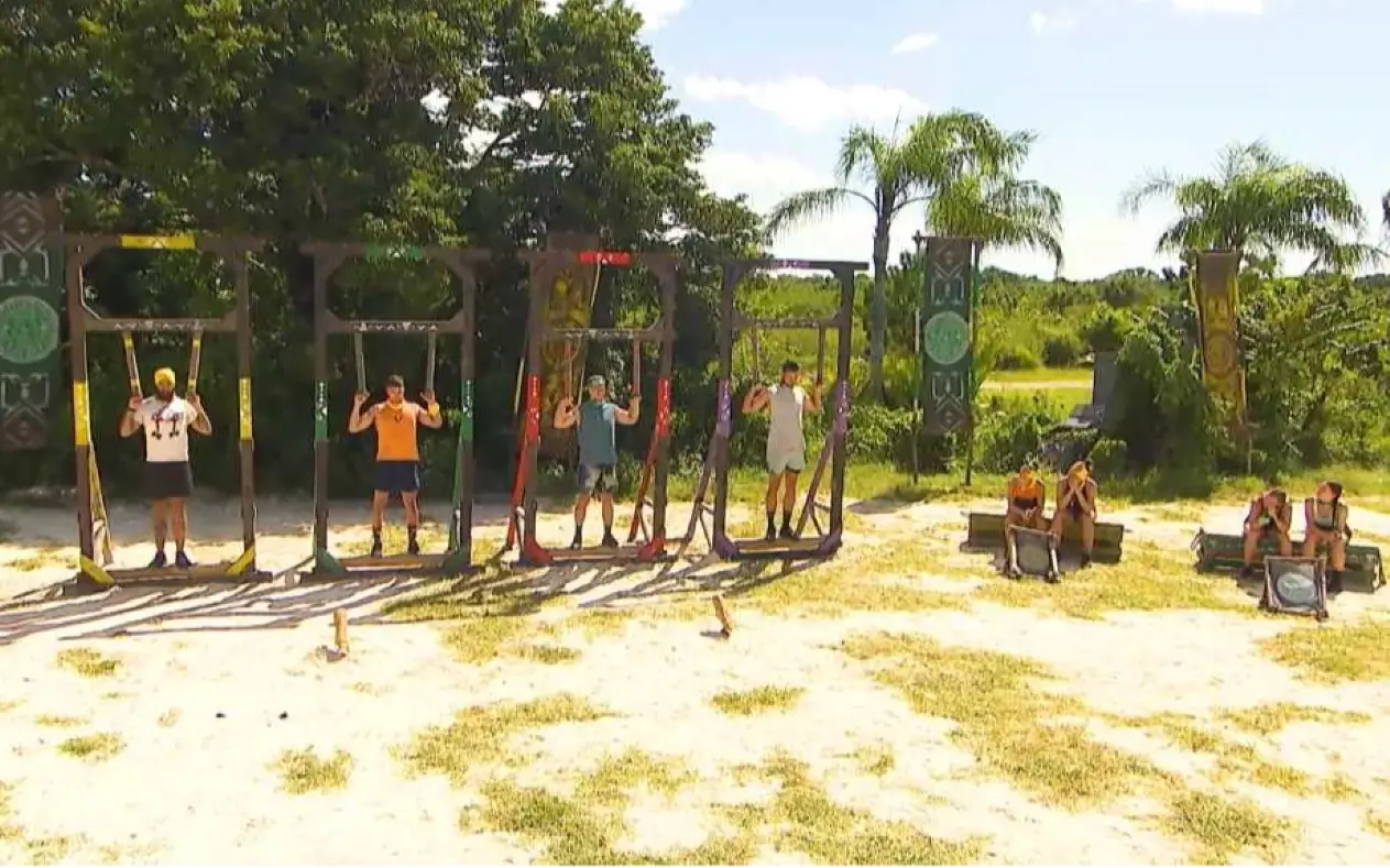 Četiri takmičara u Survivor izazovu izdržljivosti drže se za drvene okvire, dok ostali posmatraju sa strane u tropskom okruženju