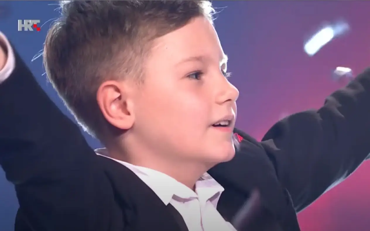 Dečak u svečanom odelu slavi pobedu u muzičkom takmičenju "The Voice Kids", dok konfete padaju oko njega.