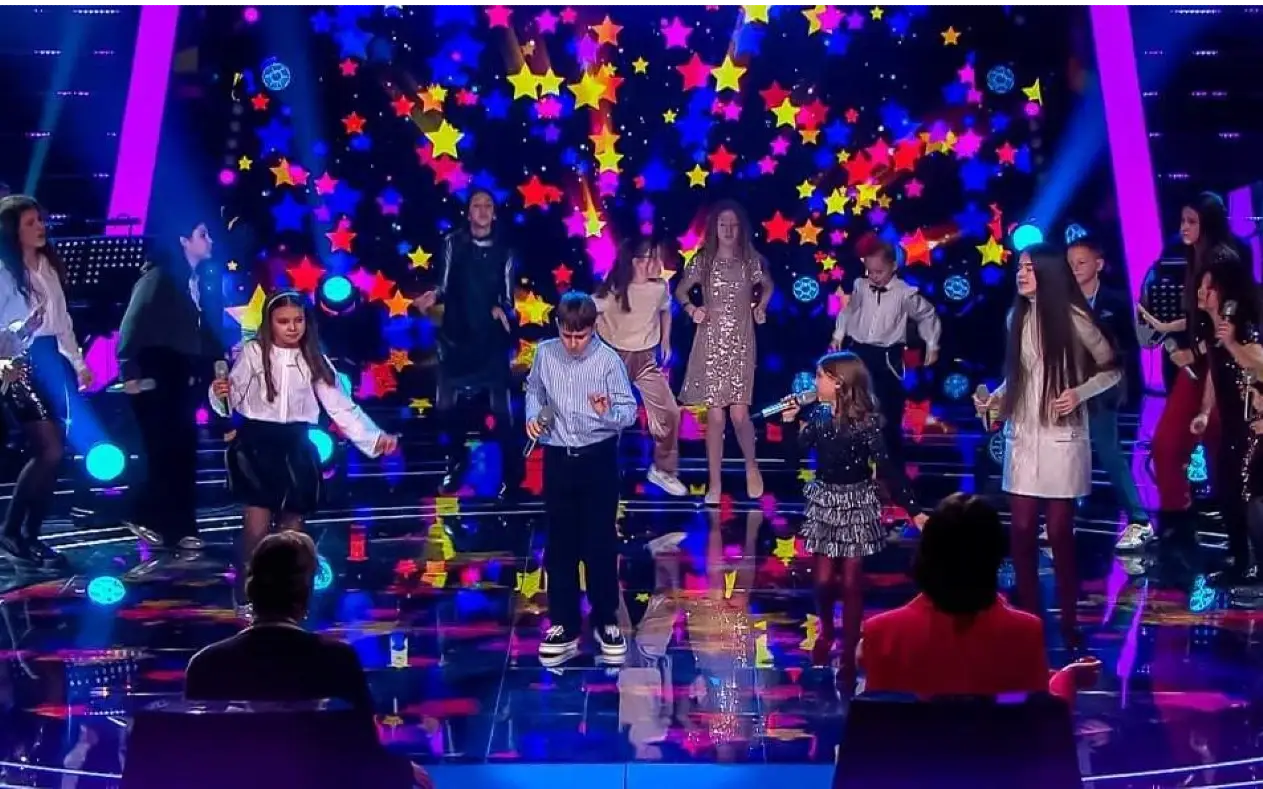 Scena šou programa "The Voice Kids" sa talentovanom decom koja pevaju i plešu uz šarenu pozadinu sa zvezdama.