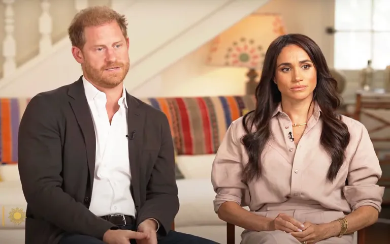 Meghan Markle i princ Harry sede jedno pored drugog u intervjuu; Hari nosi crni sako i belu košulju, dok je Megan u bež odeći, oboje izgledaju pažljivo i ozbiljno.