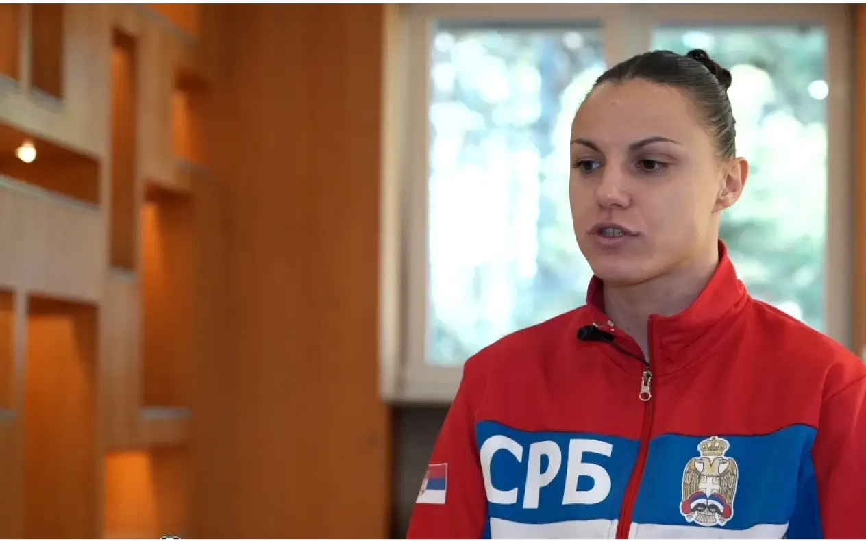 Anđela Branković, srpska sportistkinja, nosi crveno-plavi trenerku sa grbom Srbije dok daje intervju u zatvorenom prostoru sa drvenim enterijerom.