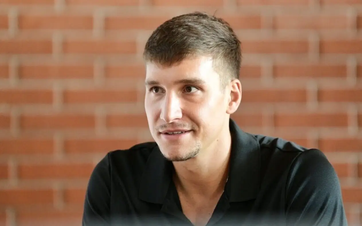 Bogdan Bogdanović, srpski košarkaš, nasmejan dok sedi ispred zida od crvene cigle, obučen u crnu Adidas majicu sa logom njegovog košarkaškog kampa.