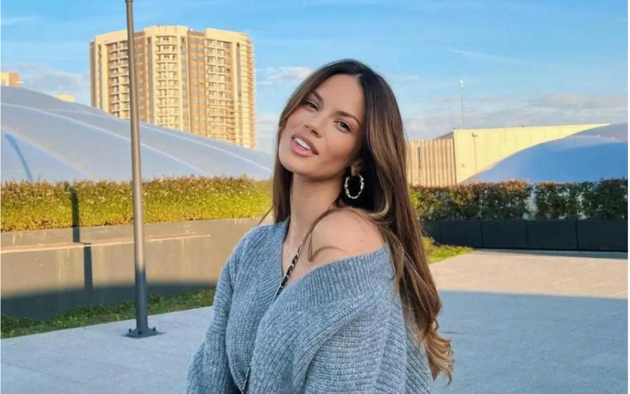 Nasmejana devojka sa dugom smeđom kosom u sivom džemperu pozira na otvorenom, sa zgradama u pozadini