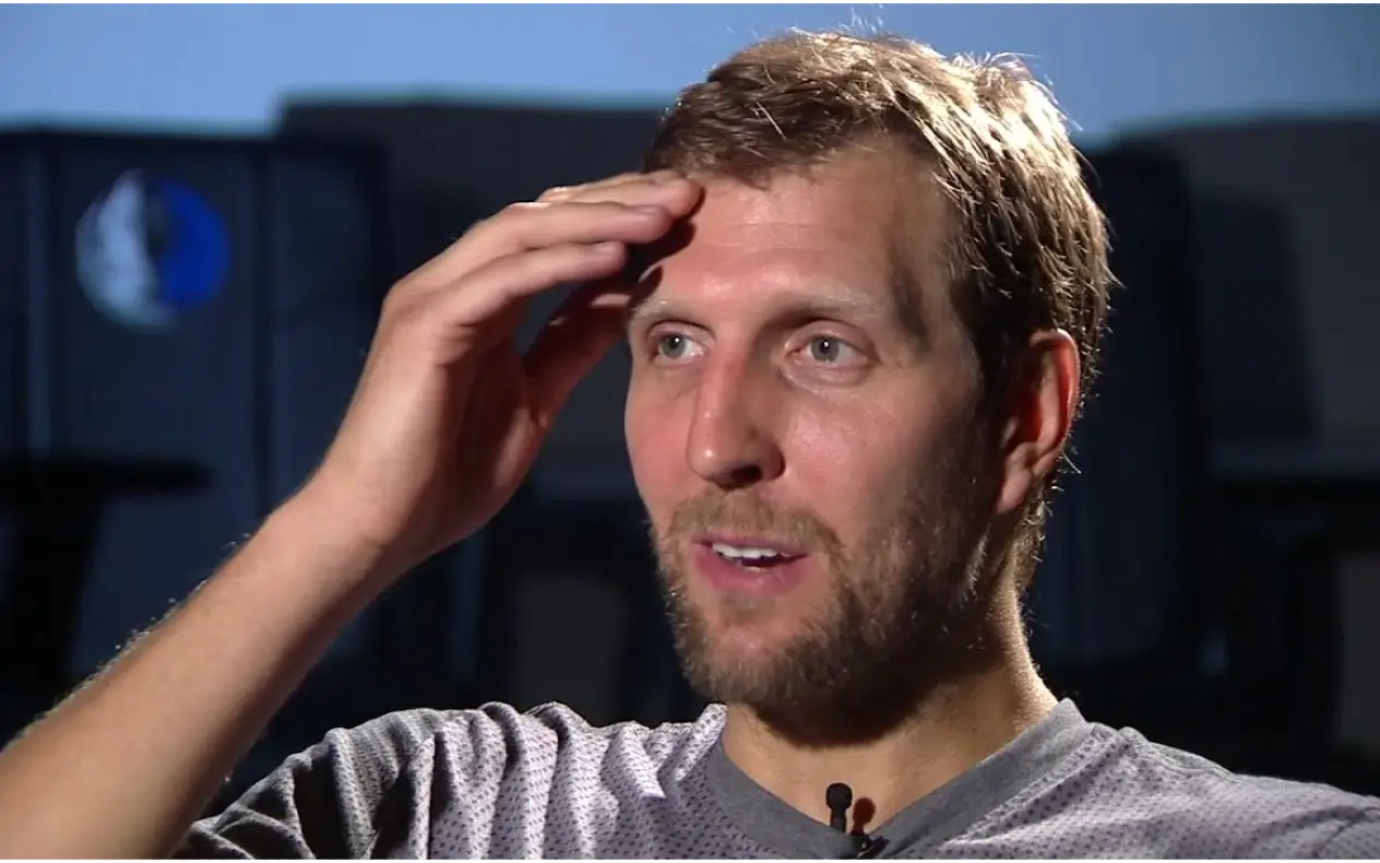 Dirk Nowitzki, bivši nemački košarkaš, nasmejan tokom intervjua, sa rukom na čelu, u pozadini sedišta sa logotipom Dallas Mavericksa.