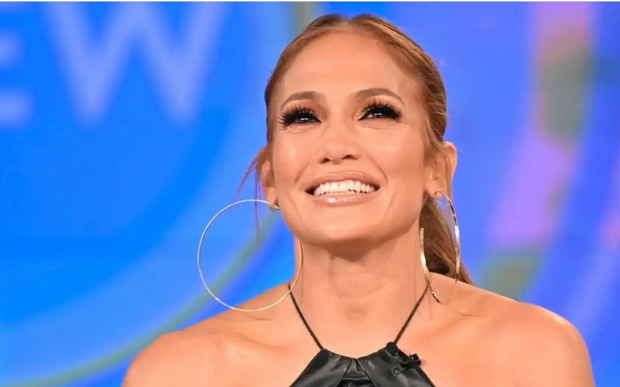 Jennifer Lopez sa širokim osmehom, u crnoj haljini i velikim okruglim minđušama, tokom televizijskog intervjua.