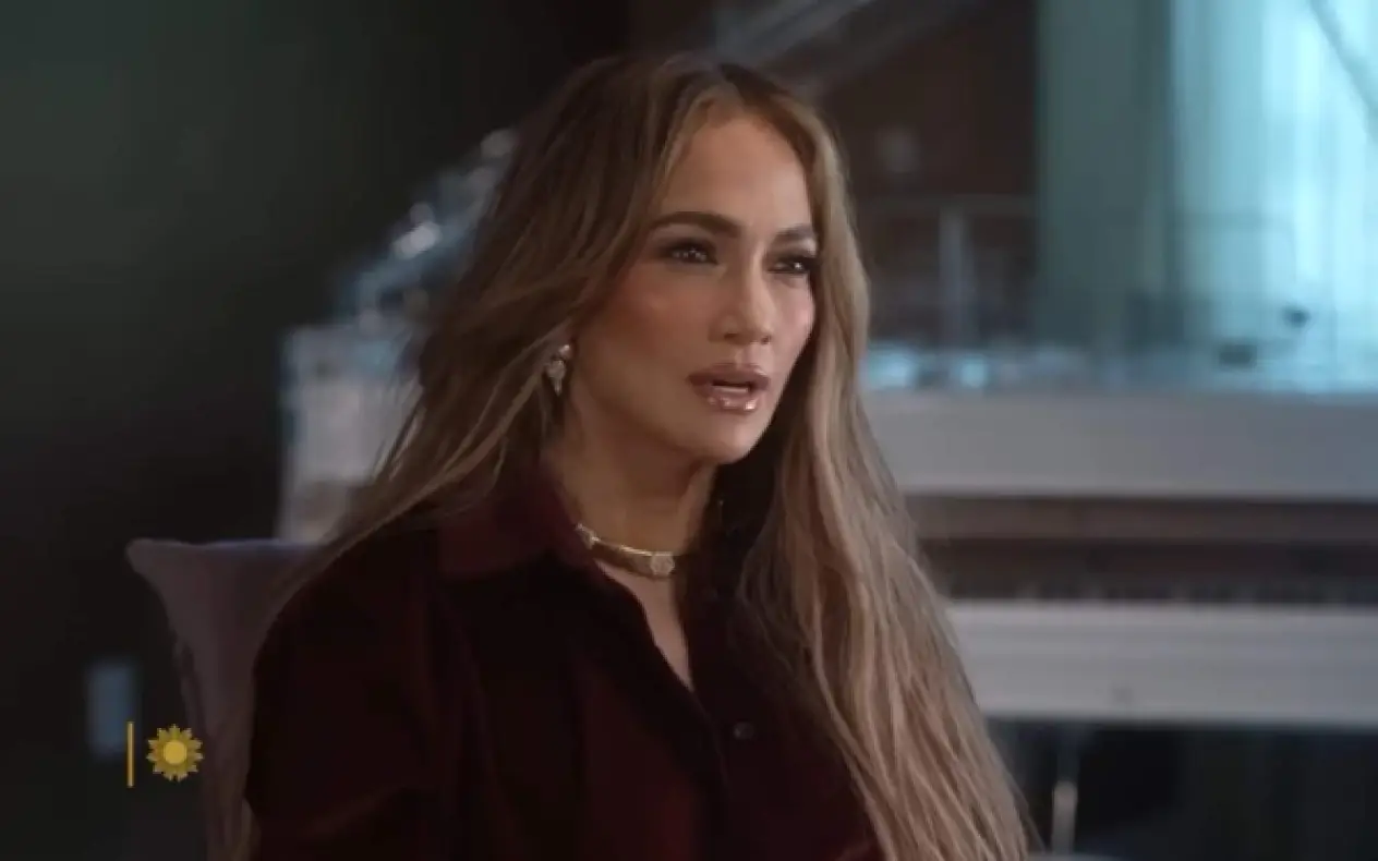 Jennifer Lopez sa dugom talasastom kosom i tamnim naočarima za sunce, obučena u tamni sako, pozira u osvetljenom luksuznom holu sa dekorativnim drvećem u pozadini.