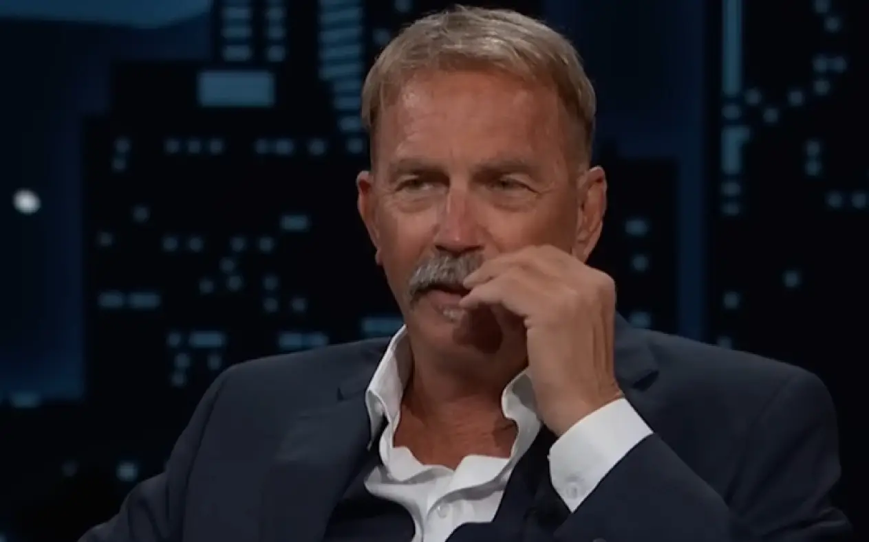Kevin Coster u teget odelu i beloj košulji dok jednom rukom dira brkove tokom gostovanja u tv emisiji