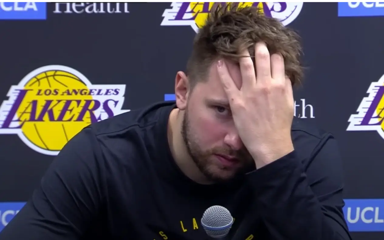 Luka Dončić sedi na konferenciji za medije, držeći ruku na čelu, sa logotipom Los Angeles Lakersa u pozadini.