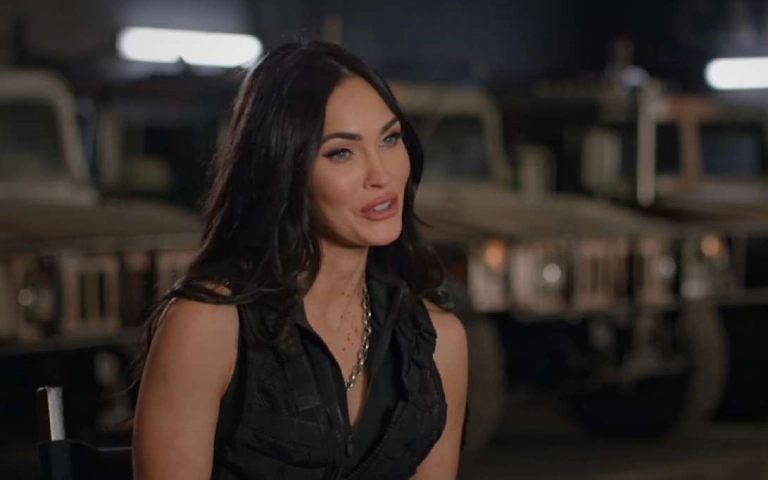 Megan Fox sedi ispred vojnih vozila, obučena u crno i govori u intervju okruženju