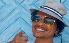 Bruno Mars sa naočarima za sunce i šeširom, nasmejan pred kamerom. Bruno Mars sa naočarima za sunce i šeširom, pokazuje prstom ka kameri u opuštenom ambijentu