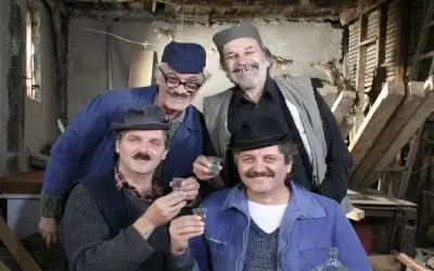 Četiri muškarca u radničkoj odeći i kapama, sede u staroj radionici i nazdravljaju čašicama rakije, smešeći se.