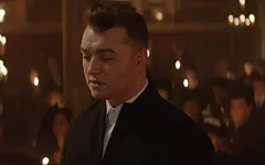 Sam Smiith u formalnom odelu u ambijentu sa svećama, zamišljenog izraza lica. Sam Smiith u tamnom odelu stoji sa zatvorenim očima, okružen ljudima koji drže sveće