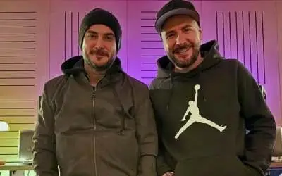 Muzički duo DNK u studiju, obučeni u crne dukseve i kape, nasmejani dok poziraju ispred osvetljenog zida sa ljubičastim tonovima.