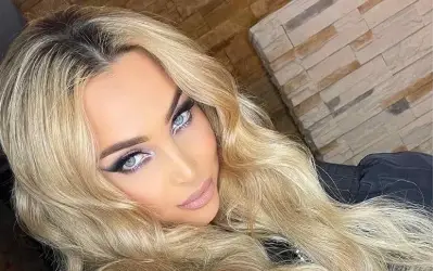 Pevačica Maya Berović sa dugom plavom kosom i naglašenim šminkom, pozira ispred zidne dekoracije od kamena.