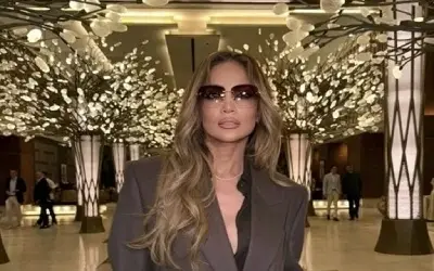 Jennifer Lopez sa dugom talasastom kosom i tamnim naočarima za sunce, obučena u tamni sako, pozira u osvetljenom luksuznom holu sa dekorativnim drvećem u pozadini.