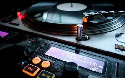 Bliski prikaz DJ opreme, uključujući gramofon sa pločom i digitalni mikser sa osvetljenim ekranom i dugmadima.