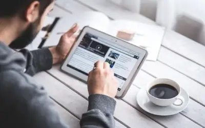 Muškarac drži tablet i čita najnovije vesti uz šoljicu kafe na drvenom stolu.