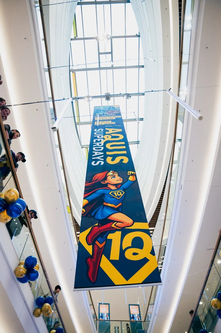 Großes Jubiläumsbanner der Aquis Superdays mit Superheldin und der Zahl 10 hängt im Atrium des Aquis Plaza