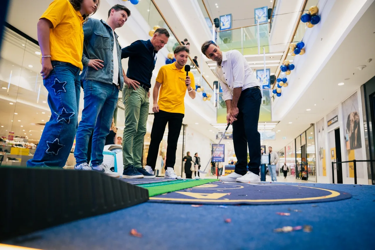 Fynn Frings moderiert mit Mikrofon ein Minigolf-Spiel im Aquis Plaza, während ein Gast zum Schlag ansetzt