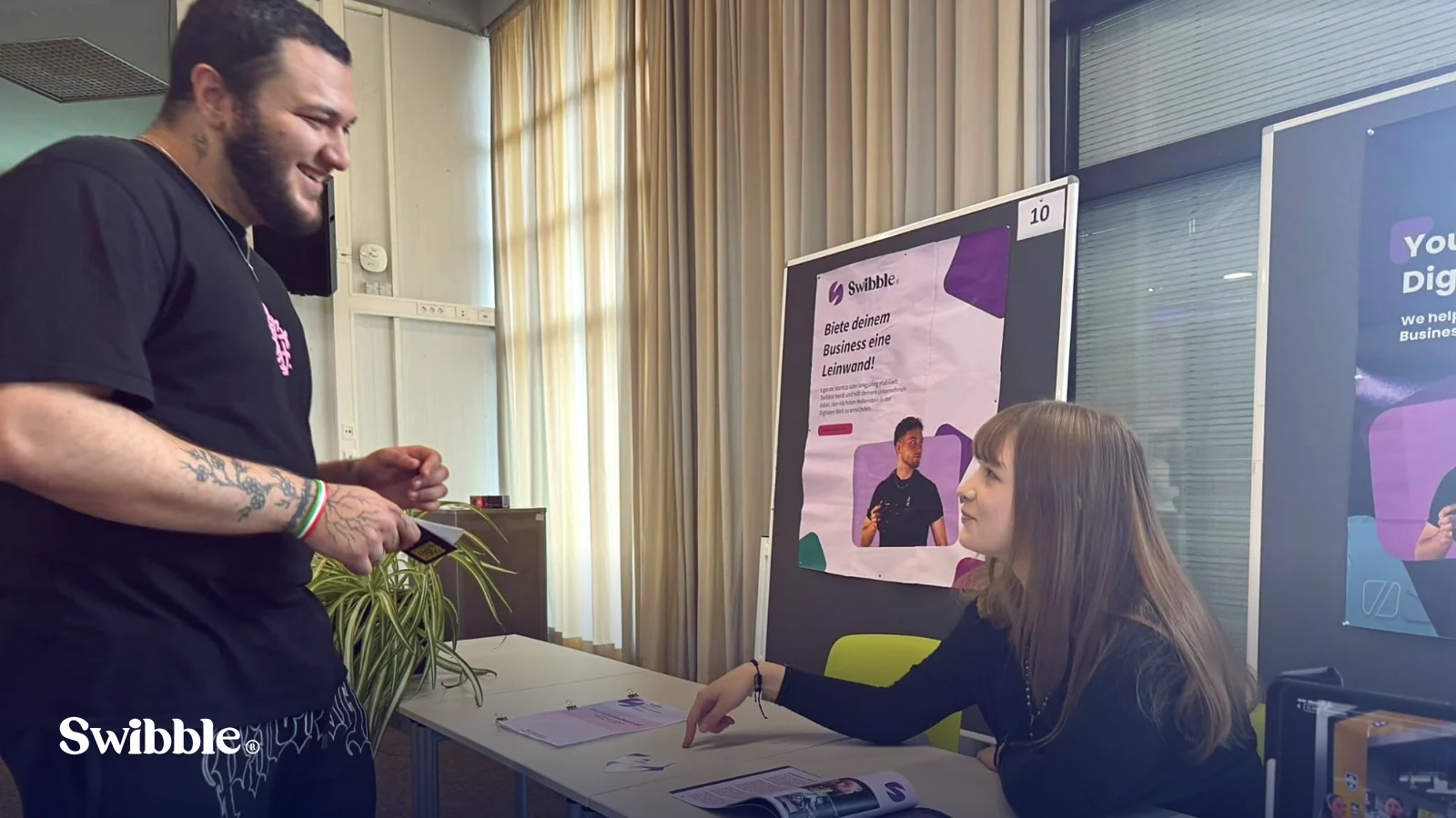 Gespräch mit einem lachenden Besucher am Swibble-Stand der Praktikumsmesse, auf dem Tisch liegen der Praktikumsbericht und Swibble-Visitenkarten